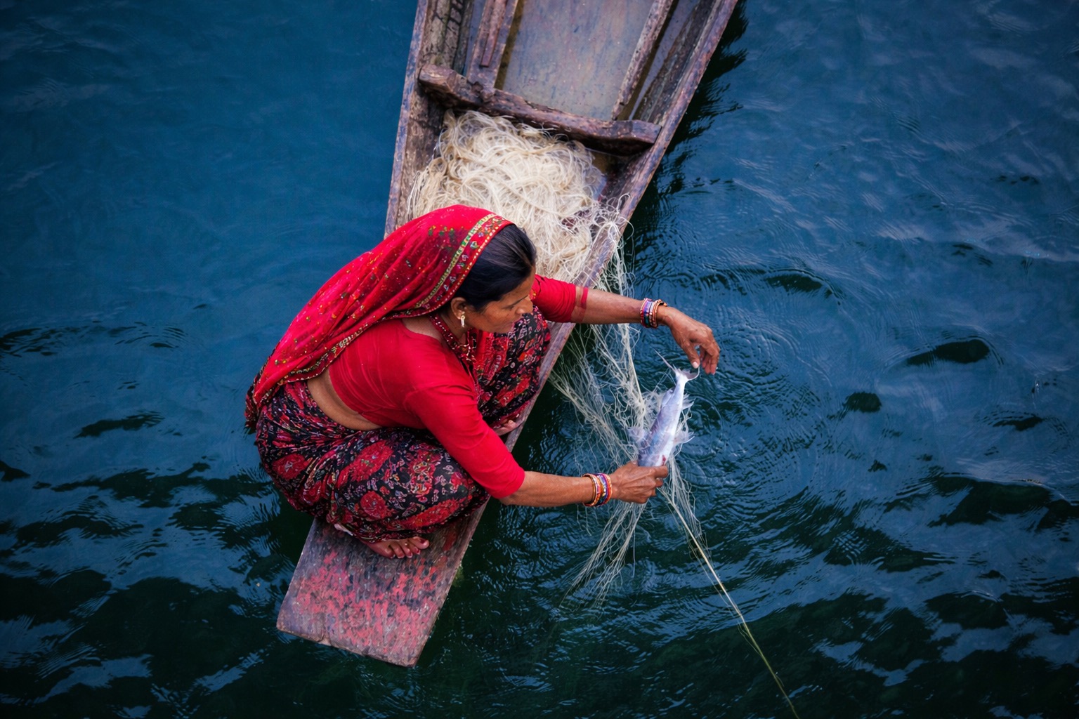 A woman crouches on a wooden boat while pulling a fish from a net.